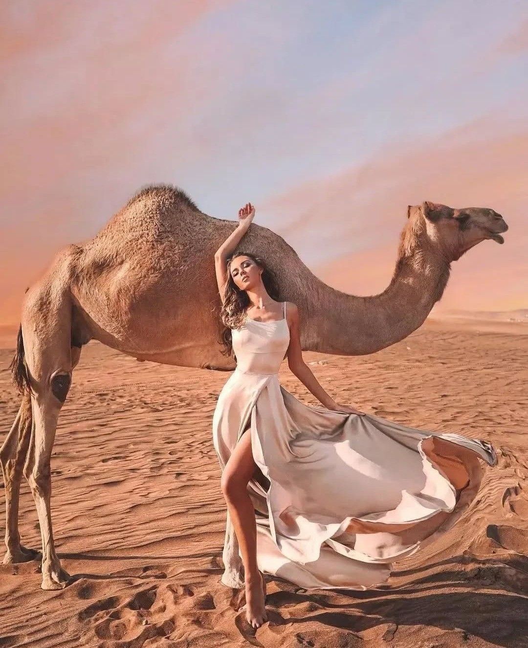 Elegant Dubai desert fashion photoshoot with model in flowing dress beside camel at sunset