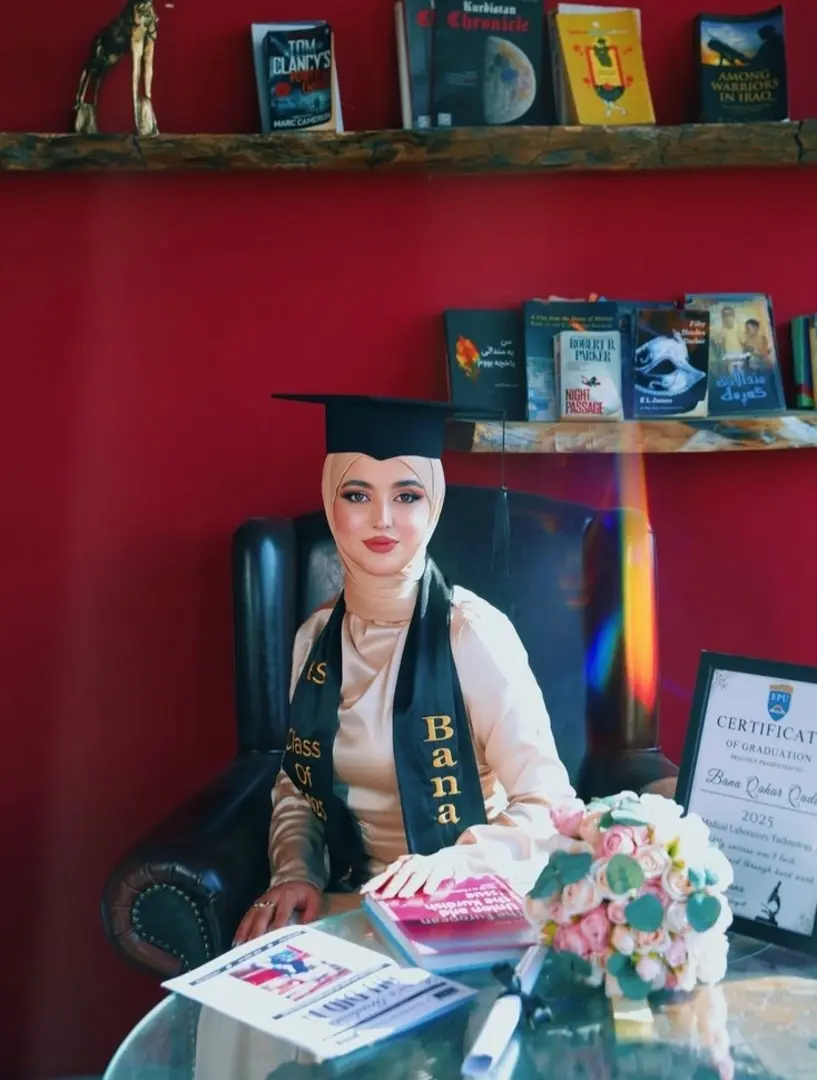 Graduate in cap and gown posing with books and a graduation certificate during an indoor graduation photoshoot in Dubai.